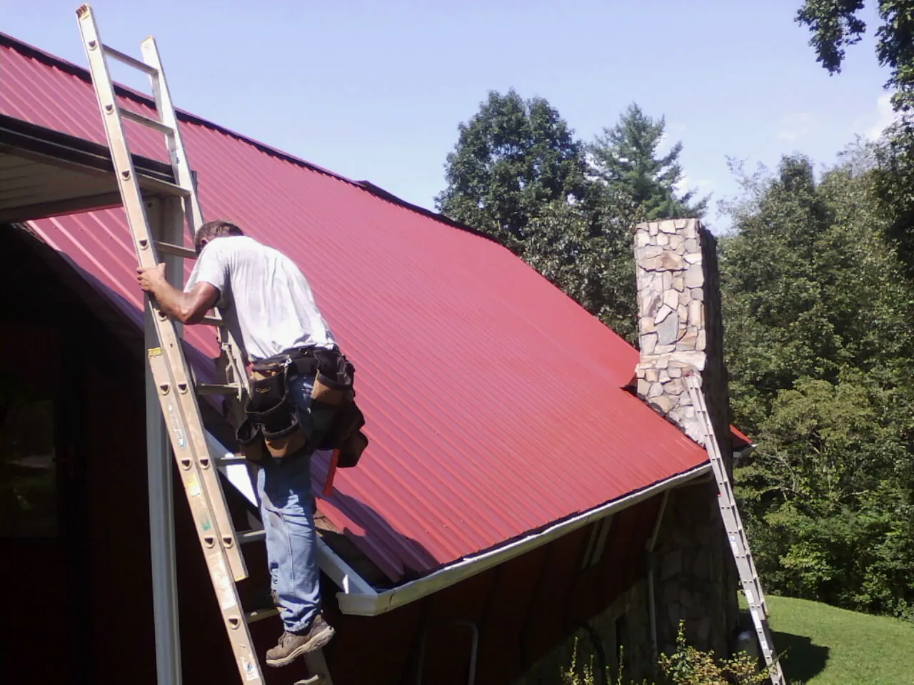 Roofer on a ladder beside a red metal roof and stone chimney — Soffit & Fascia Repair by Roofing Brothers in Havensville